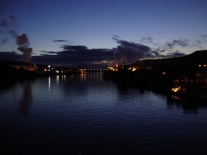 Vue de la rive gauche, au crépuscule, avec quelques bâtiments éclairés se reflétant dans l'eau du fleuve.