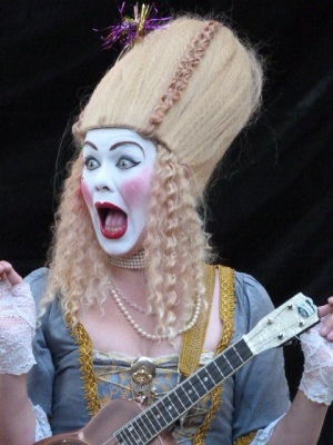 Portait expressif d'une comédienne en robe et perruque blonde, maquillée comme un clown blanc, avec son instrument de musique.